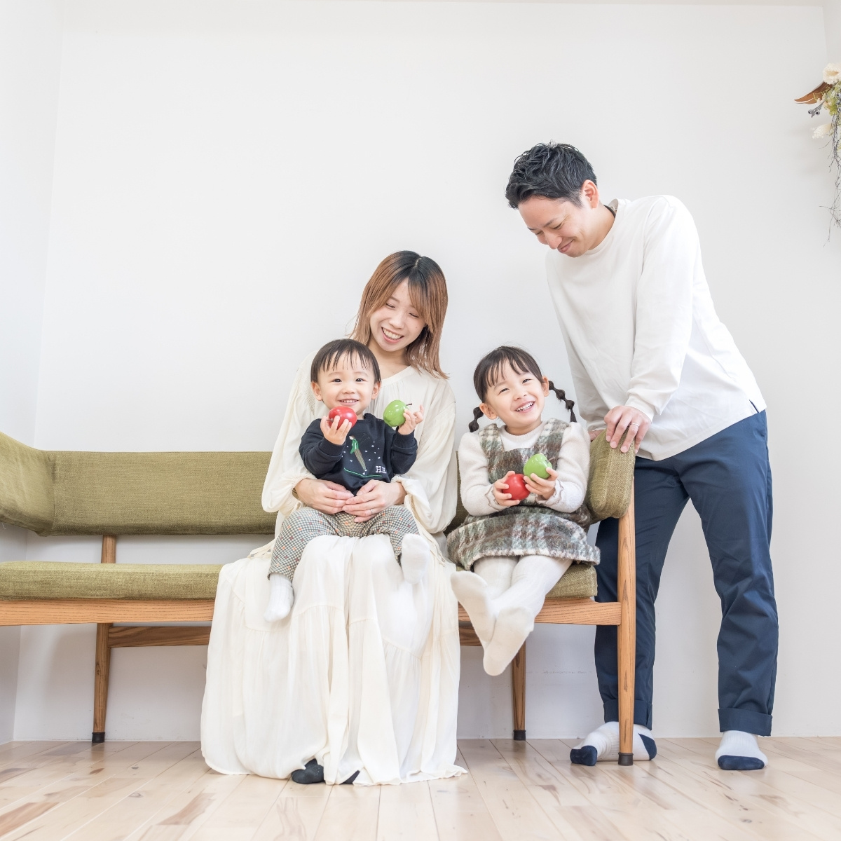 ソファに座るママとお子様2人を、パパがそばで見守る家族写真