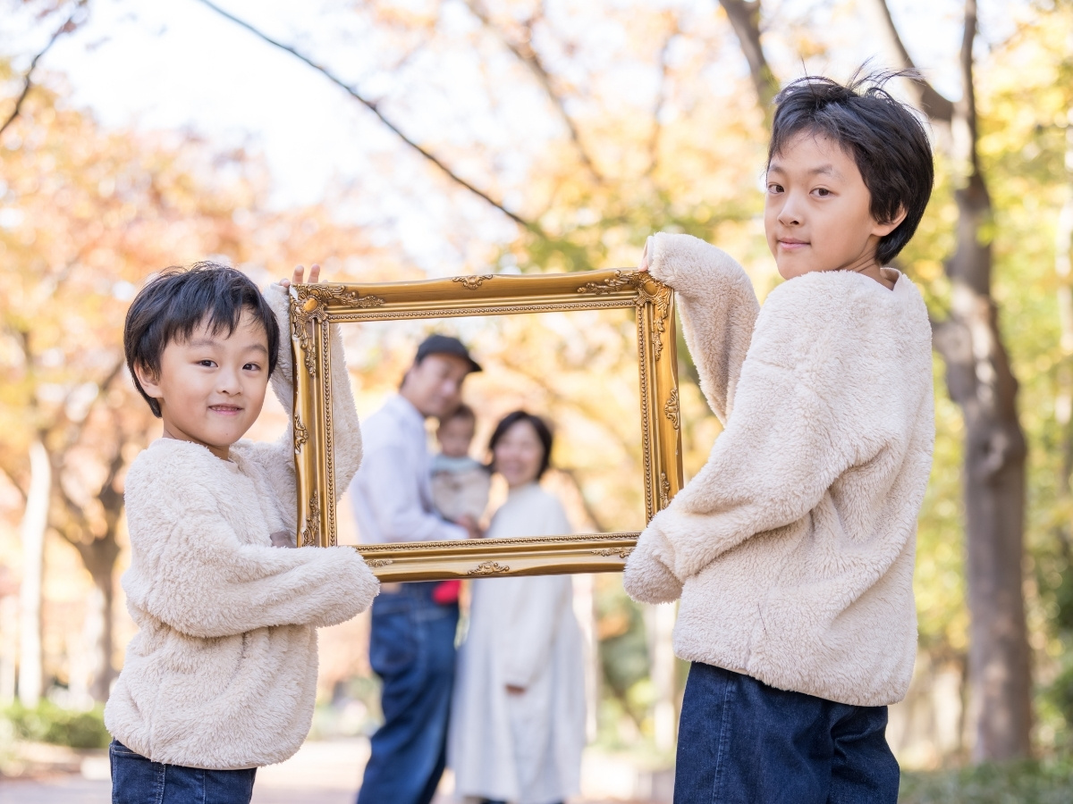 靱公園の紅葉の中で、お子様2人が持つ額縁の中にご両親と赤ちゃんを写した家族写真